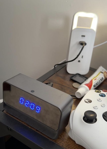 Digital clock, phone charger, and game controller on table.