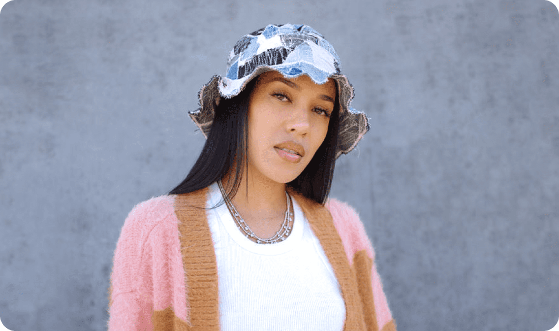 A person wearing a bucket hat and pink cardigan with brown accents