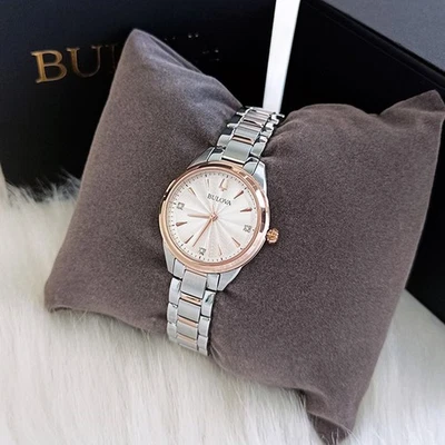 Reloj Bulova 98P200 Lady Sutton plateado con detalles en oro rosa, bisel de cristal y correa metálica estriada sobre un cojín marrón.
