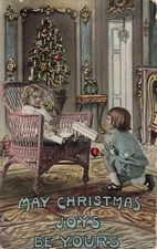 CHRISTMAS POSTCARD BOY GIVES LITTLE GIRL IN WICKER CHAIR PRESENT c1910 100825 U