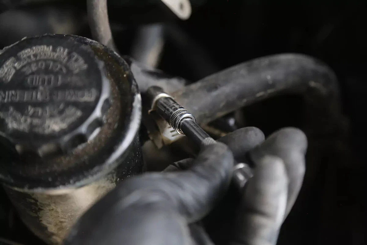 A mechanic wearing a black glove uses a small ratchet to loosen the hose clamp around the power steering fluid return line.