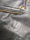 Black leather Langlitz motorcycle jacket with gold zipper, button closure, and circular gold emblem on left chest.