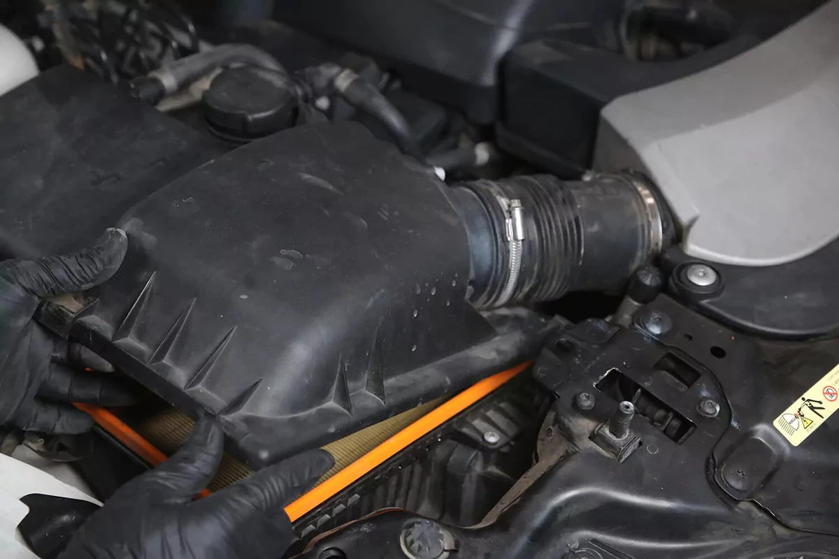 A mechanic wearing black gloves replaces the lid of the engine air filter box.