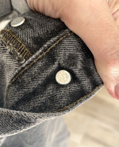Close-up of worn black Levi’s 501 XX jeans with silver rivets and button fly closure, showing metal rivets and yellow stitching.