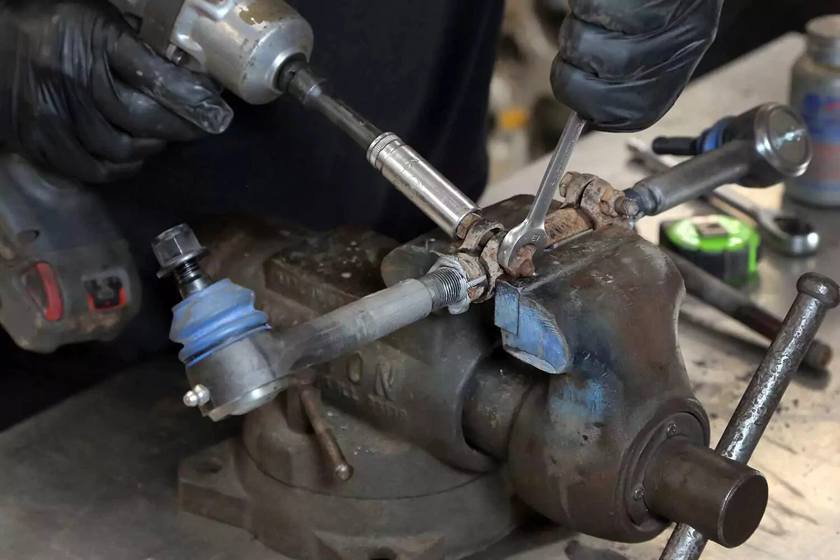 A mechanic tightens a tie rod sleeve bolt using an impact gun and an open-end wrench.