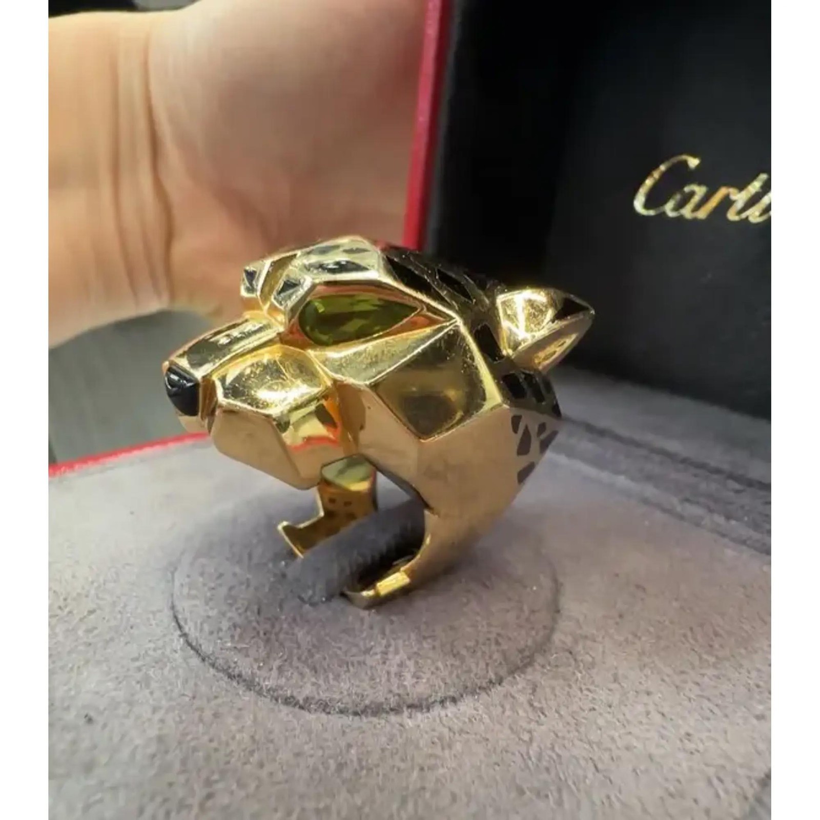 An image of a Cartier women's ring with gemstones, displayed at a close-up and slightly elevated angle. The ring, with a bold design featuring large black and green peridot gemstones, is positioned on a grey stand with the brand's name visible on a black background in the upper right corner. The yellow gold polished finish of the ring reflects the light, highlighting its intricate details and craftsmanship.