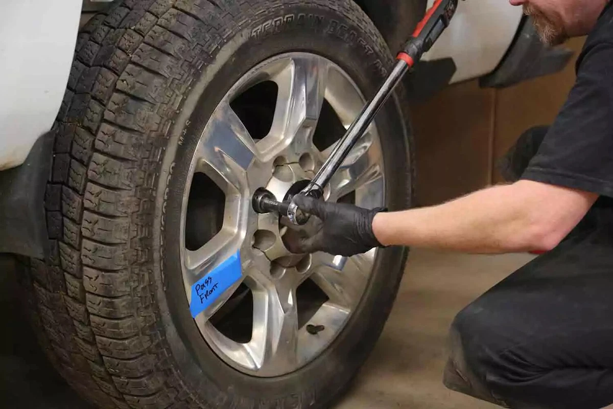 A mechanic wearing black gloves uses an electronic torque wrench with a long handle to mount a tire onto a vehicle.
