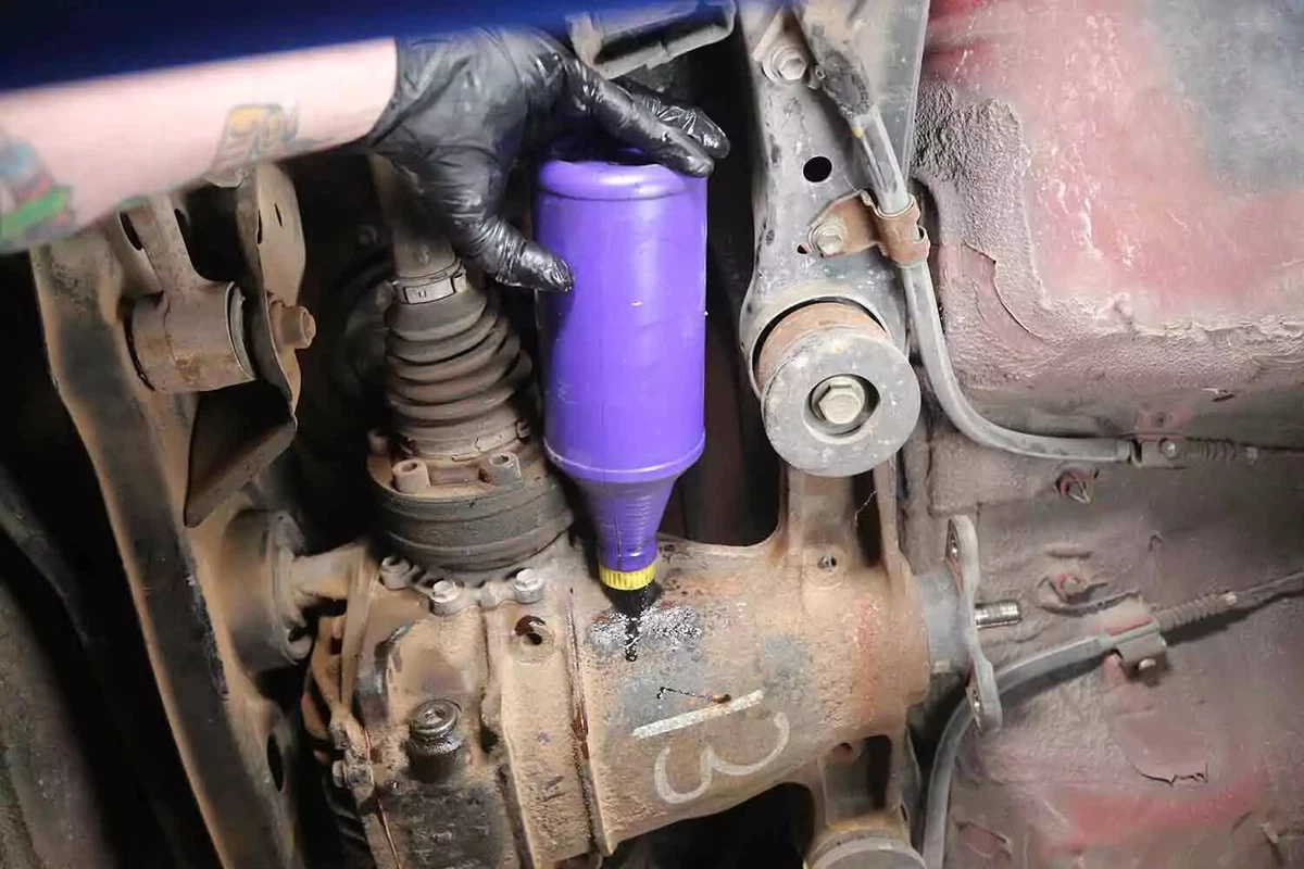 A mechanic wearing a black glove empties a bottle of differential fluid into the rear differential of a vehicle.