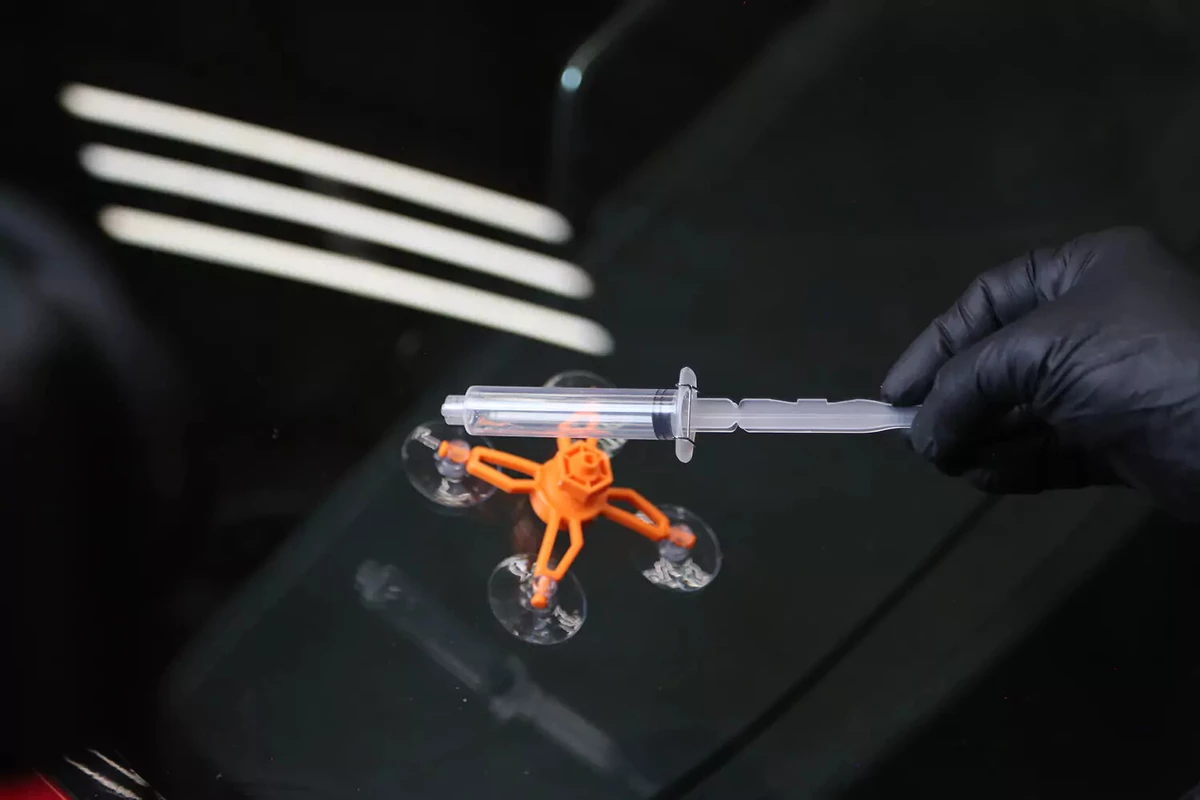 A mechanic wearing a black glove holds the syringe of a windshield repair kit over the repair jig.