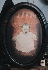 An antique convex oval wooden frame housing a creepy baby portrait — the bulging glass + faded Victorian infant image is peak 'haunted heirloom' aesthetic.