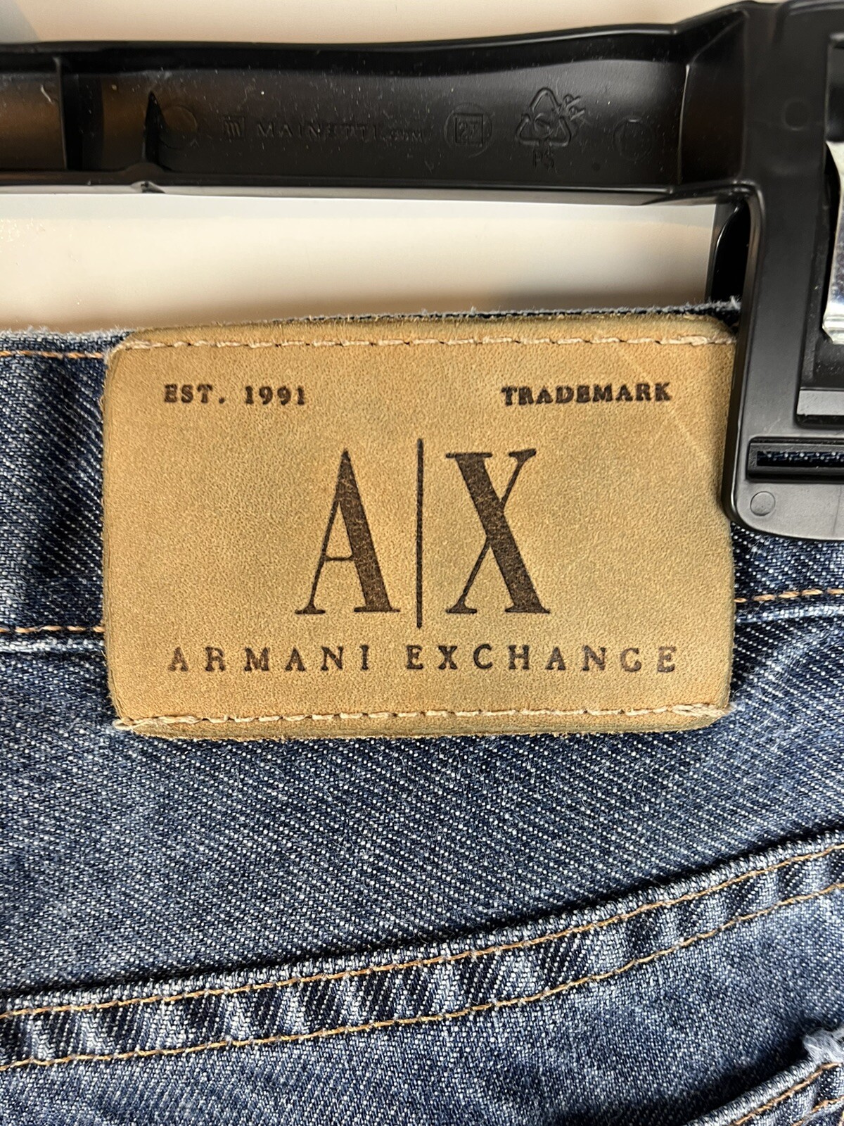 Brown leather AX Armani Exchange J40 patch with black clasp, yellow stitching, and white “AX” logo on denim fabric.