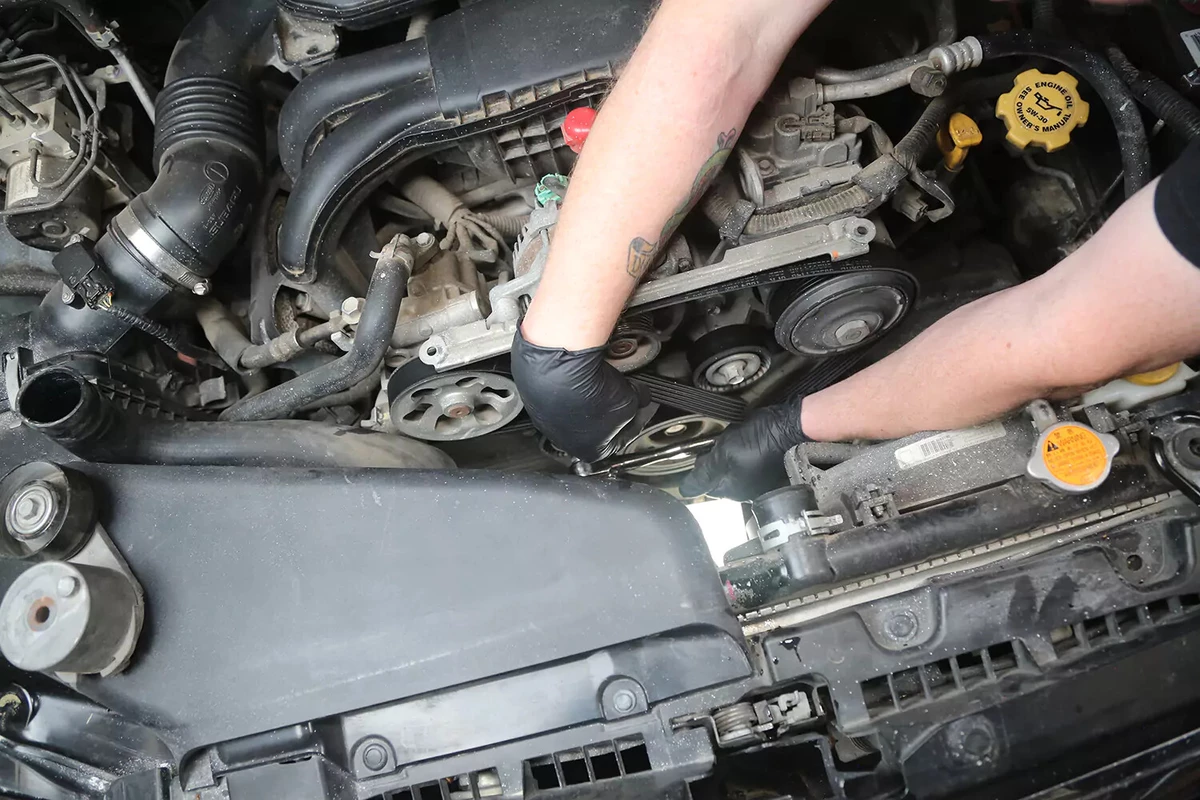 A mechanic wearing black gloves uses a ratchet to tighten the vehicle's serpentine belt tensioner.