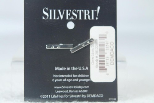 Black card with silver metal pendant shaped like a bent nail, white label reading “Demdaco” and barcode.
