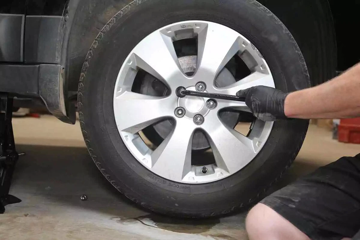 A mechanic wearing black gloves uses a tire iron to remove the lug nuts from a vehicle's tire.