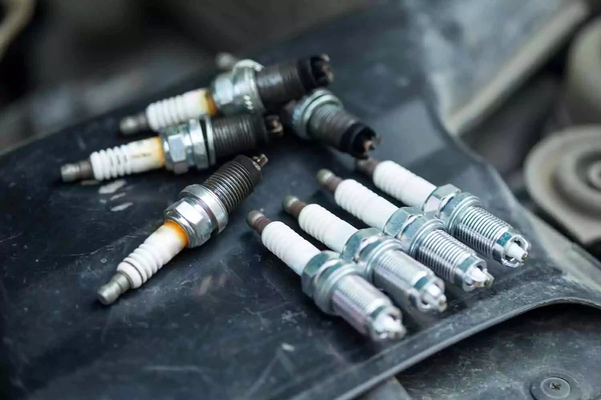 A set of four spark plugs lined up neatly, in front of a set of worn spark plugs, resting on the edge of a car’s engine compartment.