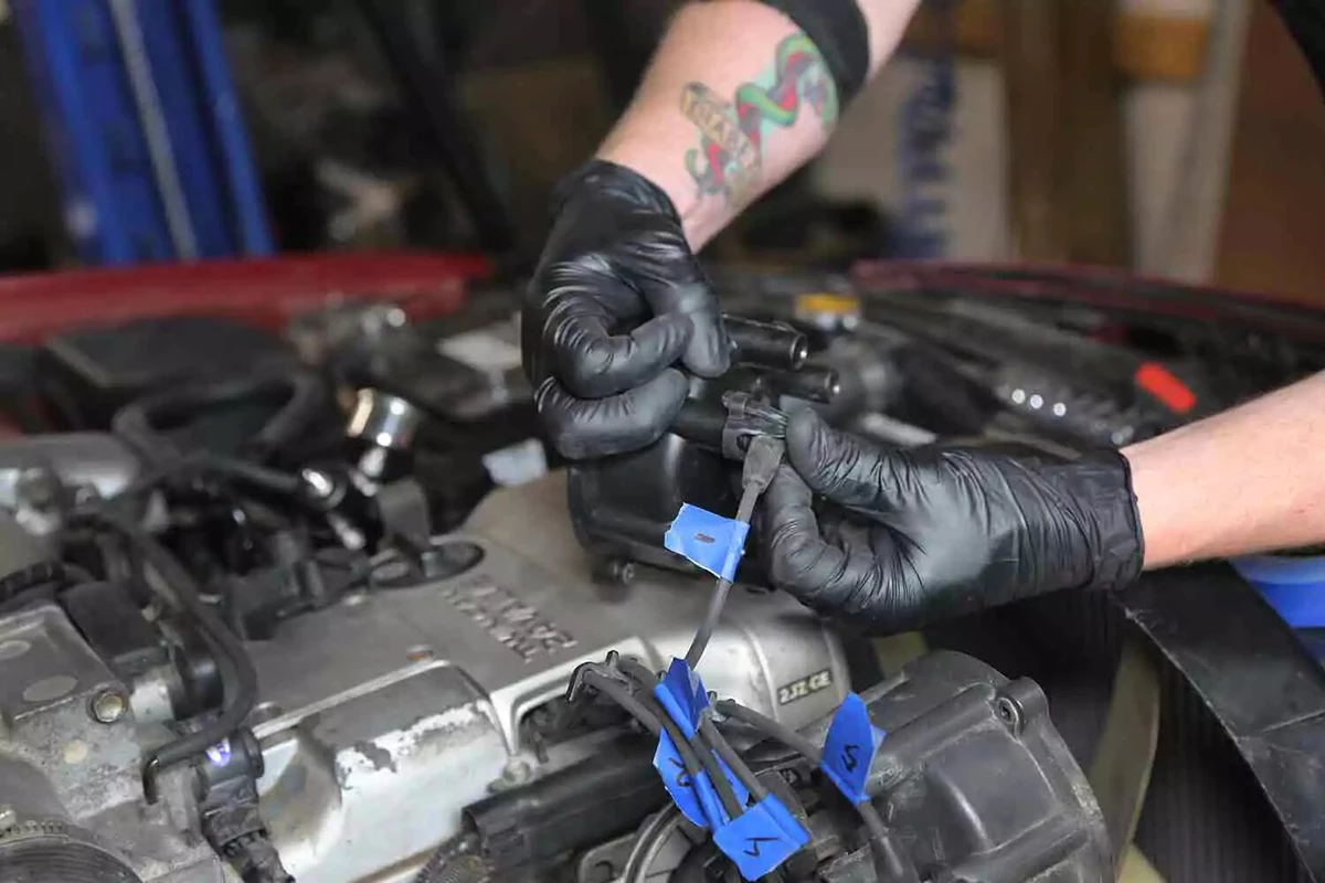 A mechanic wearing black gloves connects wires to a vehicle's distributor cap.