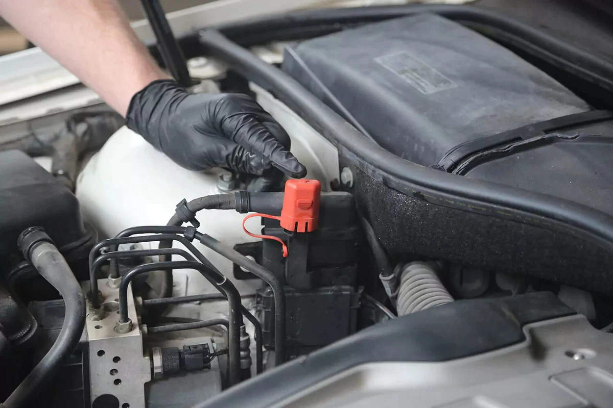 A mechanic wearing a black glove points to the red cap on the battery's positive charging point.