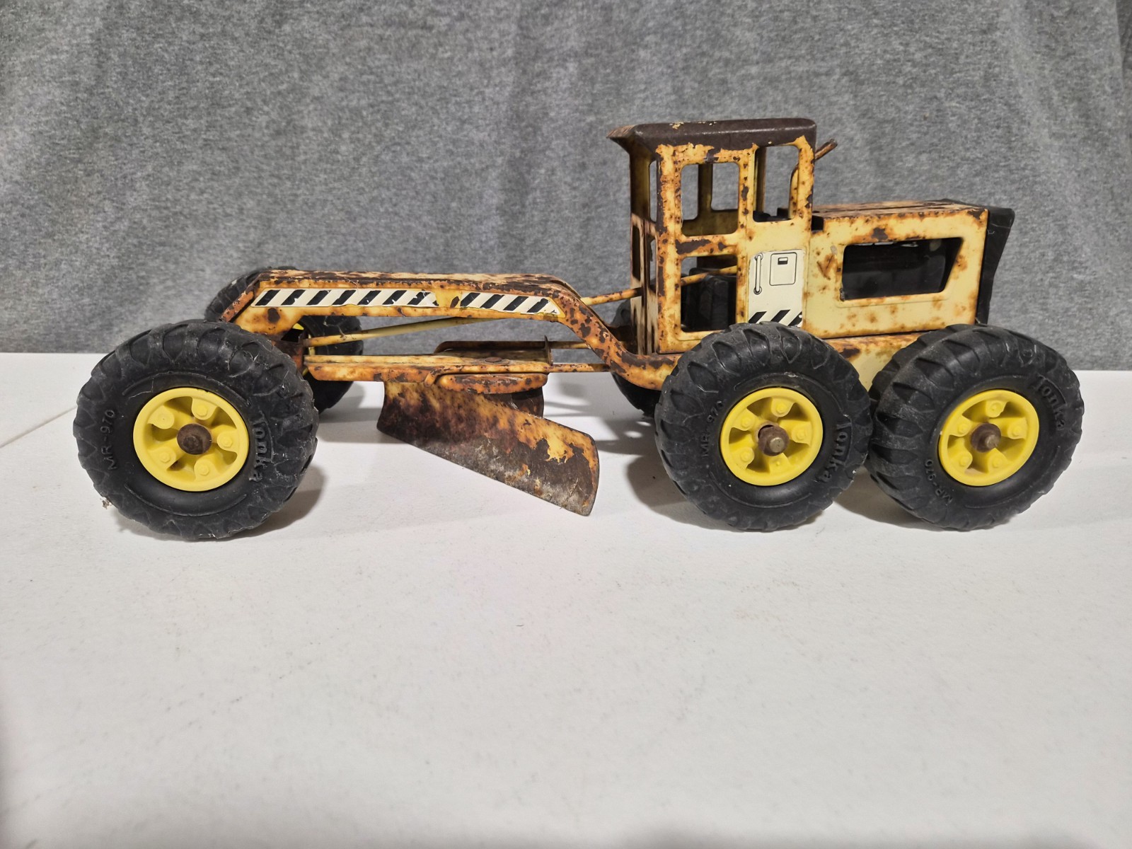 Vintage Tonka Road Grader toy, model MR-970, with worn yellow tires, black stripes, and angled blade on gray surface.