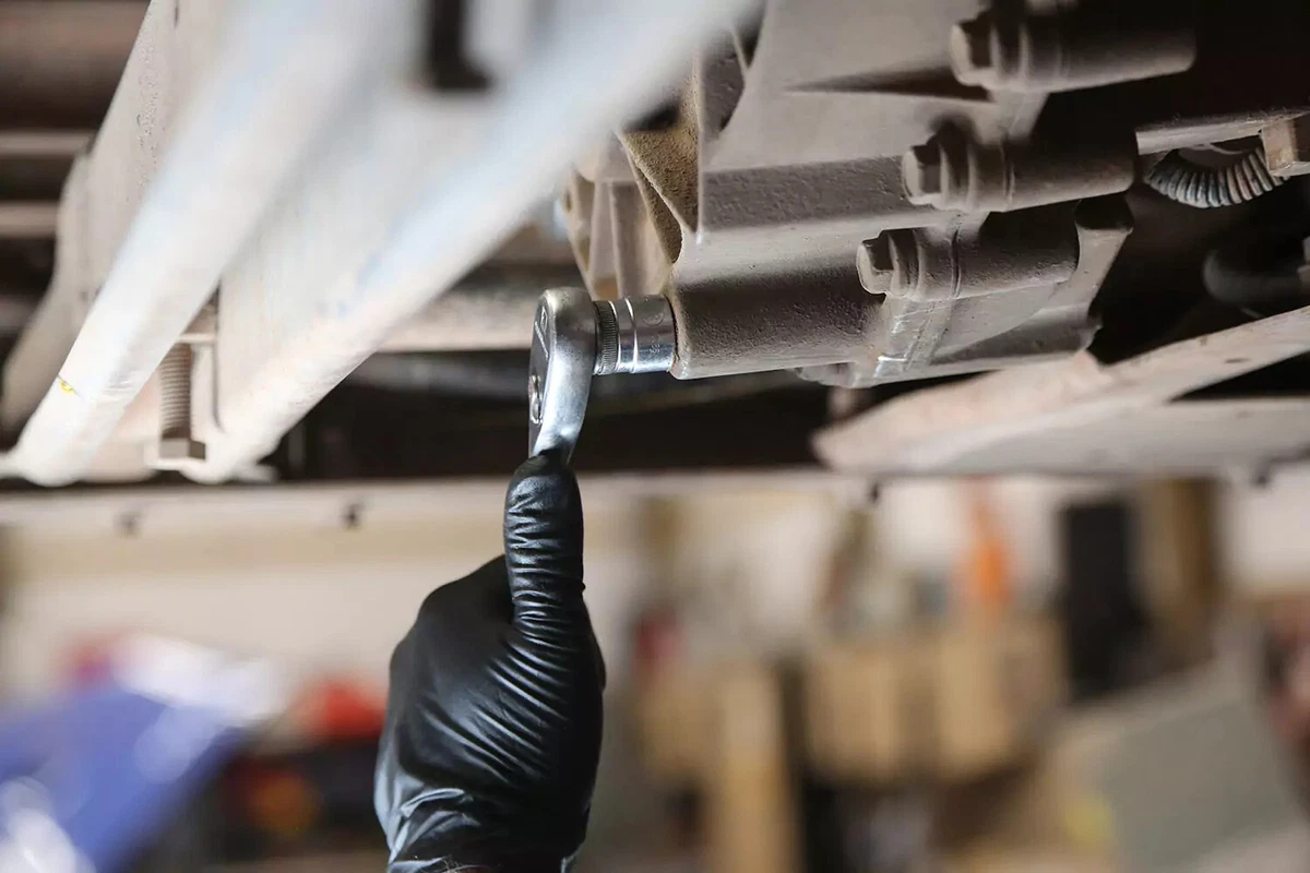 A mechanic wearing a black glove uses a ratchet to loosen the transfer case drain plug.