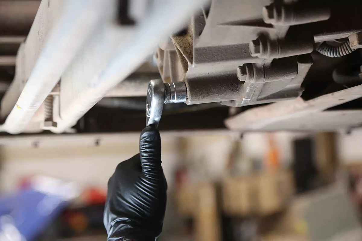 A mechanic wearing a black glove uses a ratchet to loosen the transfer case drain plug.