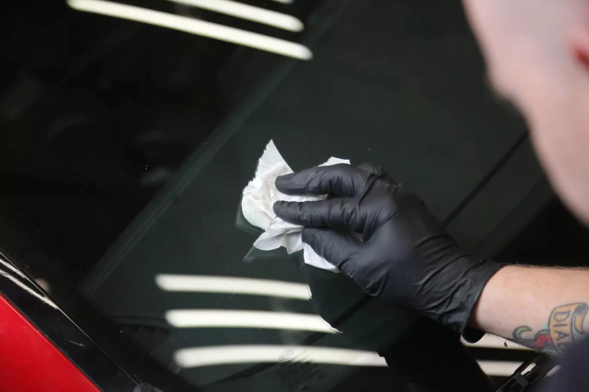A mechanic wearing a black glove uses a towel dipped in rubbing alcohol to clean the area around the windshield chip.