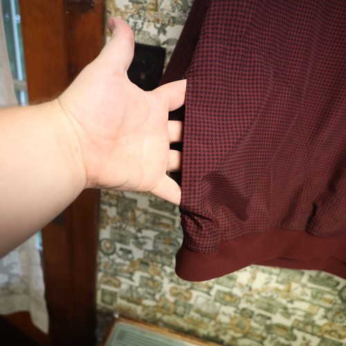 Hand touching the collar seam of a maroon VTG Zero Restriction men’s golf jacket with small black houndstooth pattern.