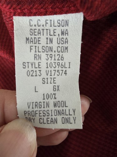 White Filson label with red and black text, size L, made in USA, dry clean only, held by hand against red fabric.