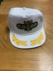 White vintage trucker hat with green and yellow wing design, “COOP” text, and tan cord across the brim.