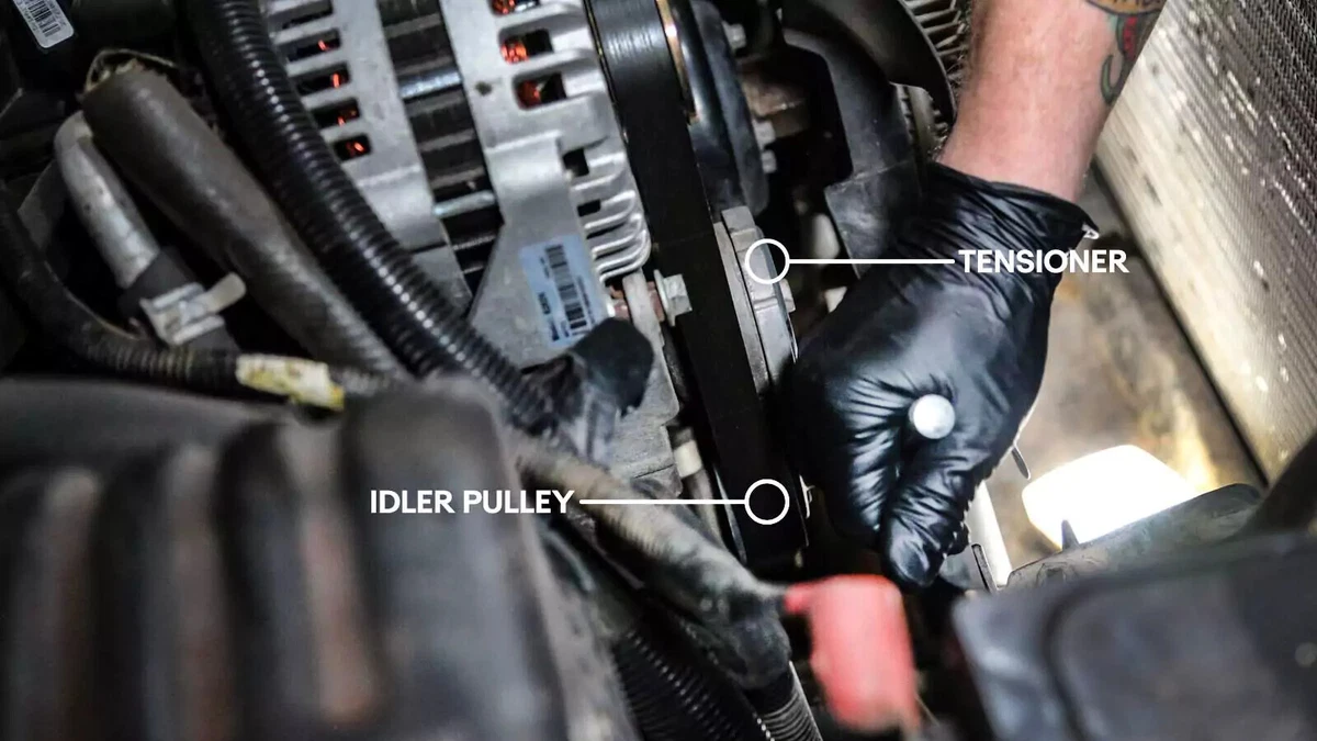 A mechanic releases the tension on the belt tensioner with a ratchet wrench. Annotations show the idler pulley and tensioner locations.
