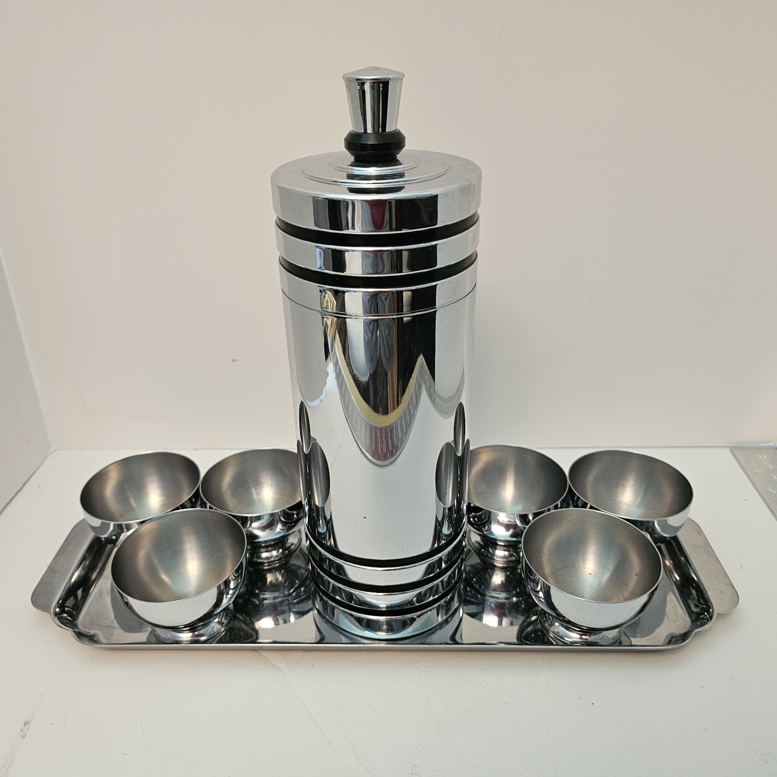 Chrome cocktail shaker with black ring details and dark stopper, surrounded by six matching chrome cordial cups on a rectangular chrome tray.