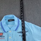Light blue Hackett London polo shirt with navy trim, buttoned collar, and embroidered Aston Martin Racing Team logo on chest.