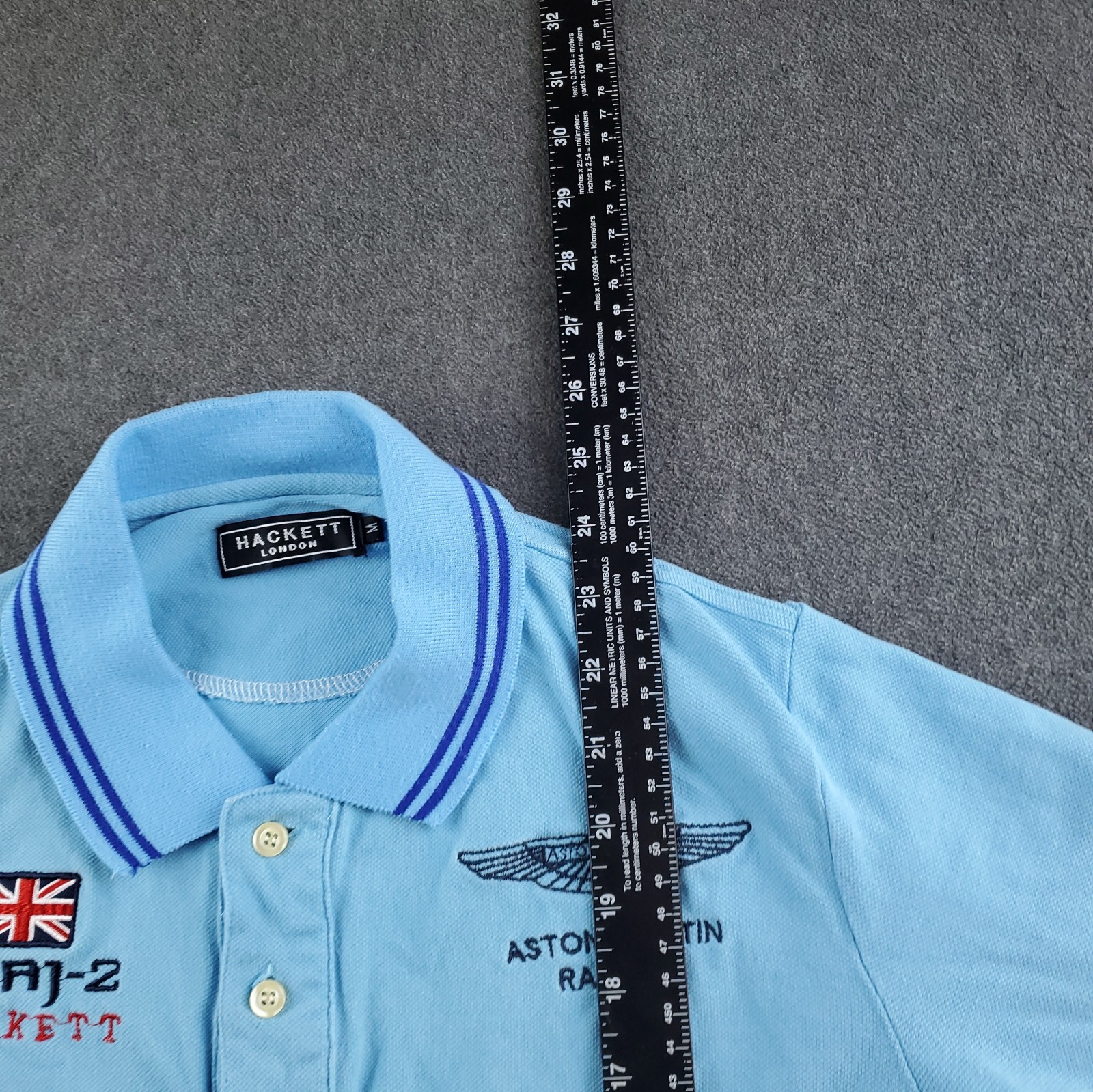 Light blue Hackett London polo shirt with navy trim, buttoned collar, and embroidered Aston Martin Racing Team logo on chest.
