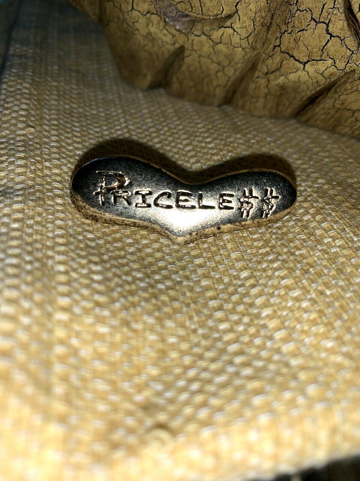 Gold-tone metal brooch with embedded text “PRICELESS” and “GENNARO MADE IN USA,” on a textured beige surface.