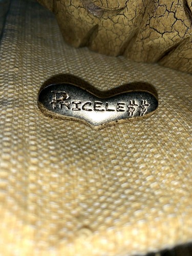 Gold-tone metal brooch with embedded text “PRICELESS” and “GENNARO MADE IN USA,” on a textured beige surface.