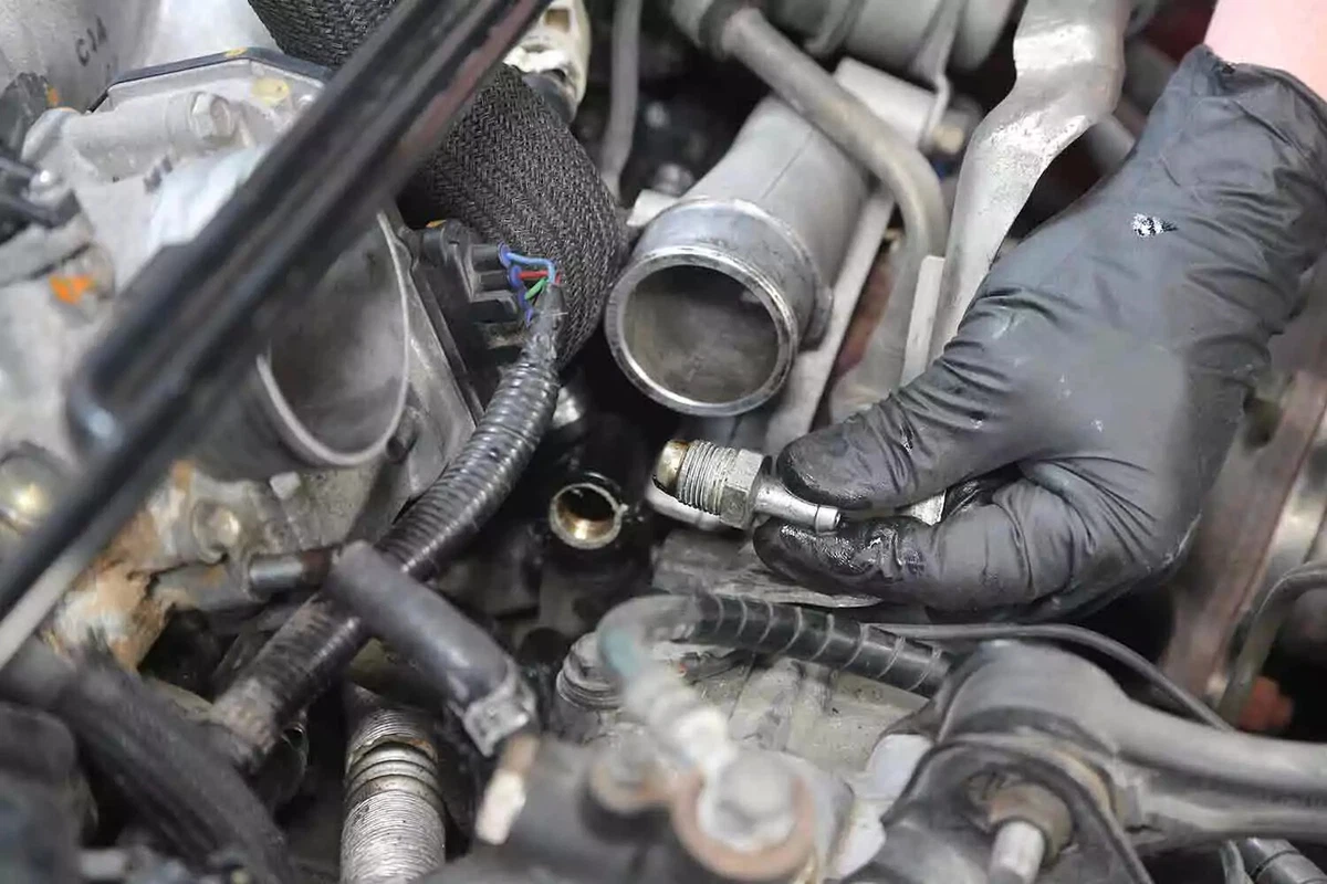 A mechanic wearing a black nitrile glove holds a PCV valve between his index finger and thumb after removing it from the engine.