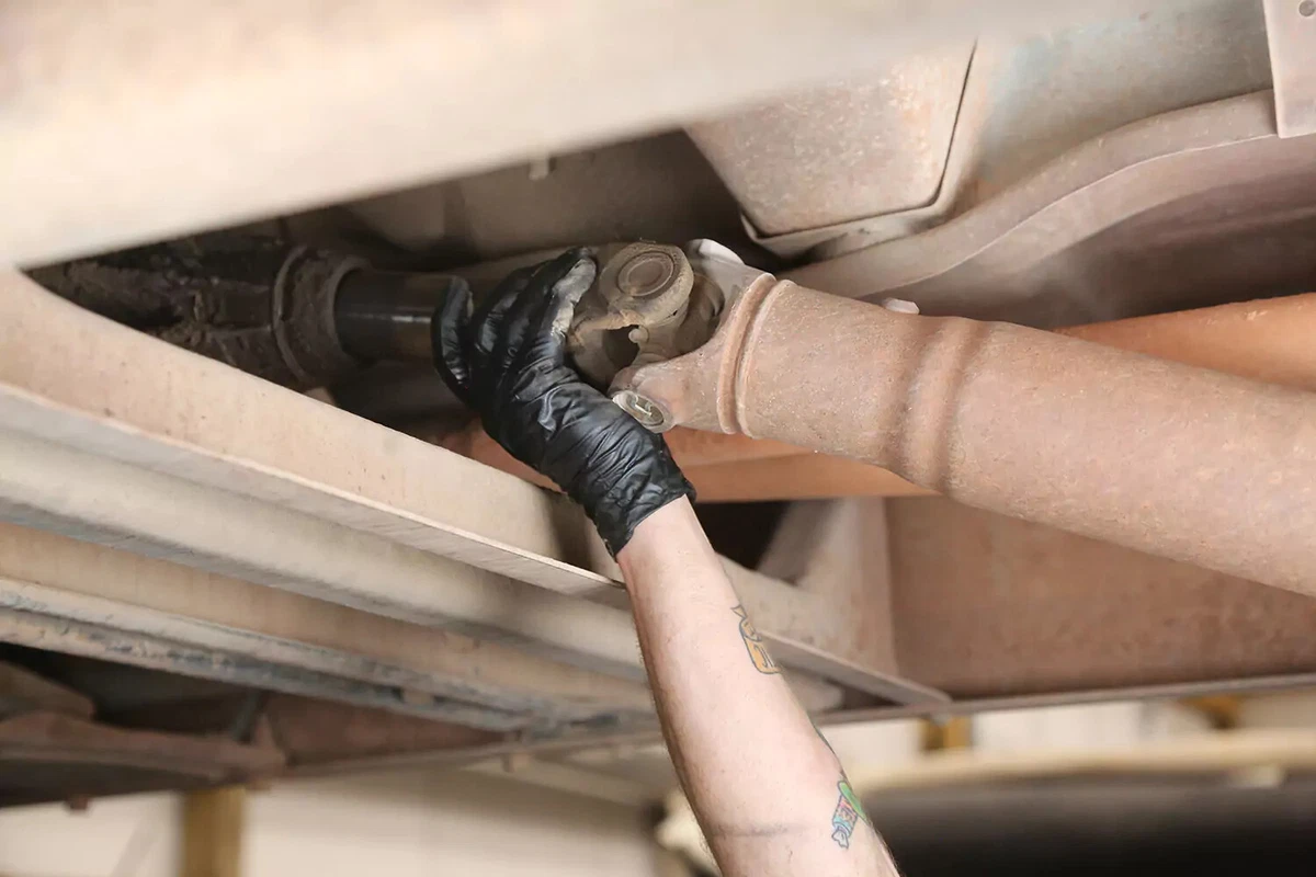 A mechanic wearing a black glove removes a vehicle's driveshaft from the transmission.