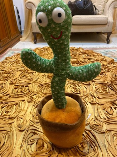 A green dancing plush cactus toy with big eyes sitting in a soft brown pot.
