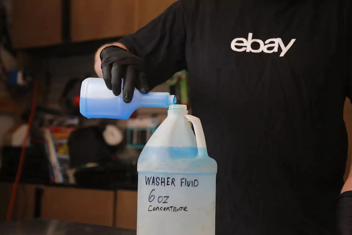 A mechanic wearing a black shirt and black glove pours blue windshield fluid concentrate into a jug of distilled water.