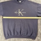 Gray sweatshirt with gold “Calvin Klein Jeans” text and logo, plus yellow measuring tape across the chest.