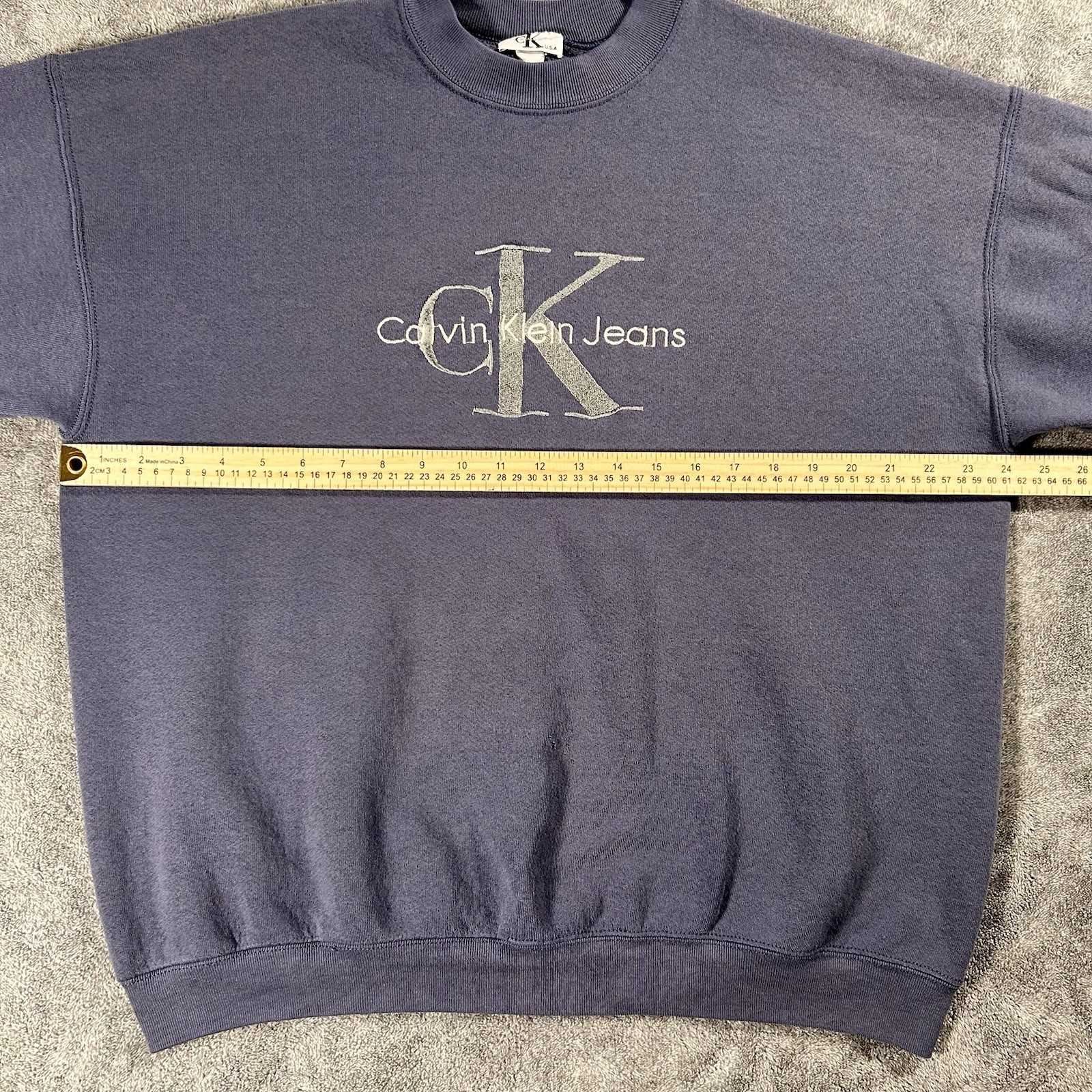 Gray sweatshirt with gold “Calvin Klein Jeans” text and logo, plus yellow measuring tape across the chest.