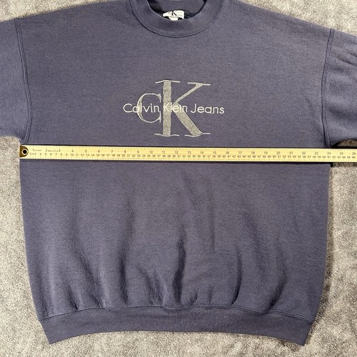 Gray sweatshirt with gold “Calvin Klein Jeans” text and logo, plus yellow measuring tape across the chest.