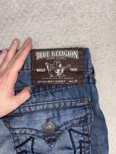 Blue distressed True Religion jeans with thick rope stitch, brown leather patch, and visible “World Tour” text.