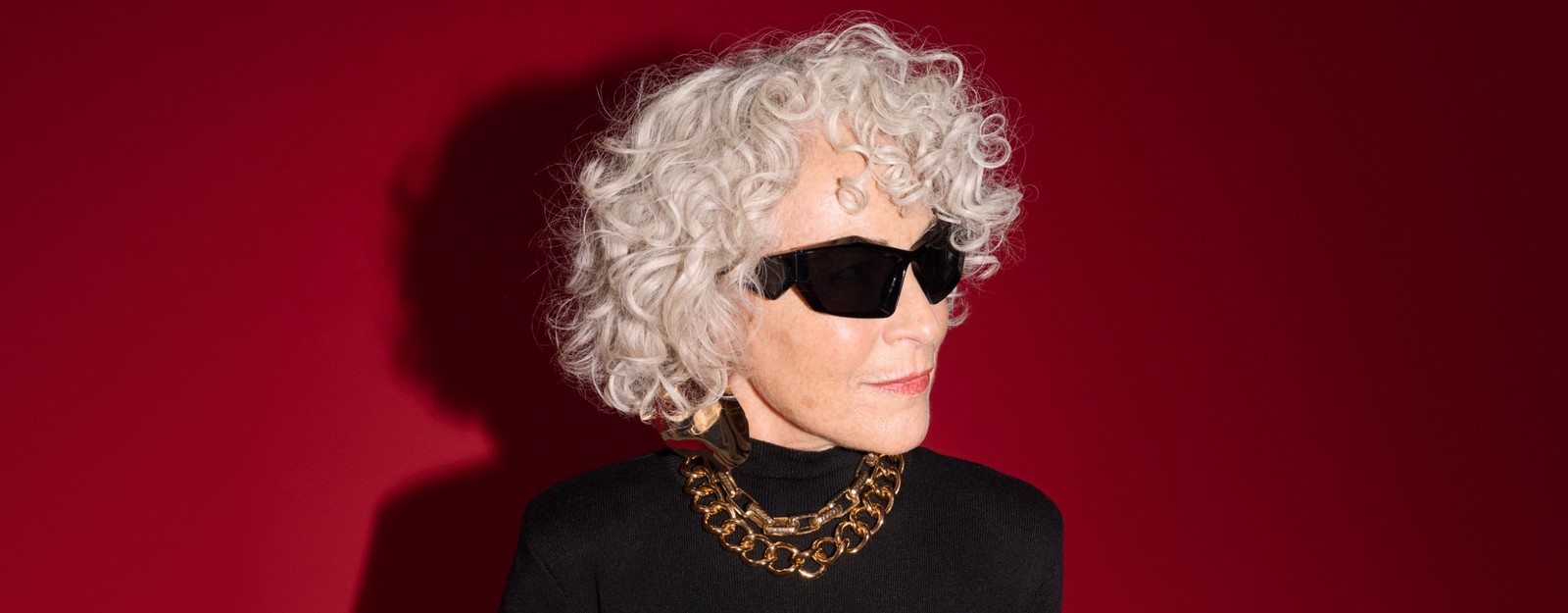 Eine Frau mit grauem lockigen Haar, einem schwarzen Rollkragenpullover, einer auffälligen goldenen Halskette und einer eckigen Sonnenbrille posiert vor einem roten Hintergrund.