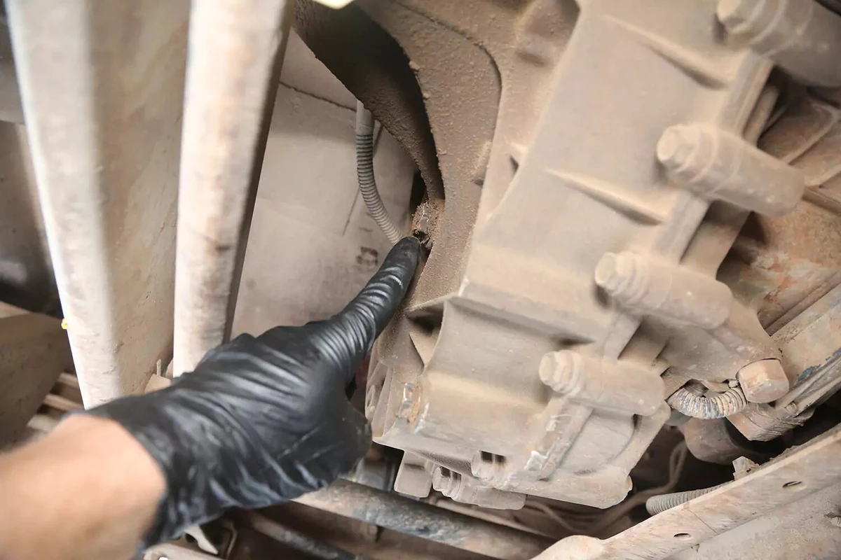 A mechanic wearing a black glove points to the fill plug of a transfer case.