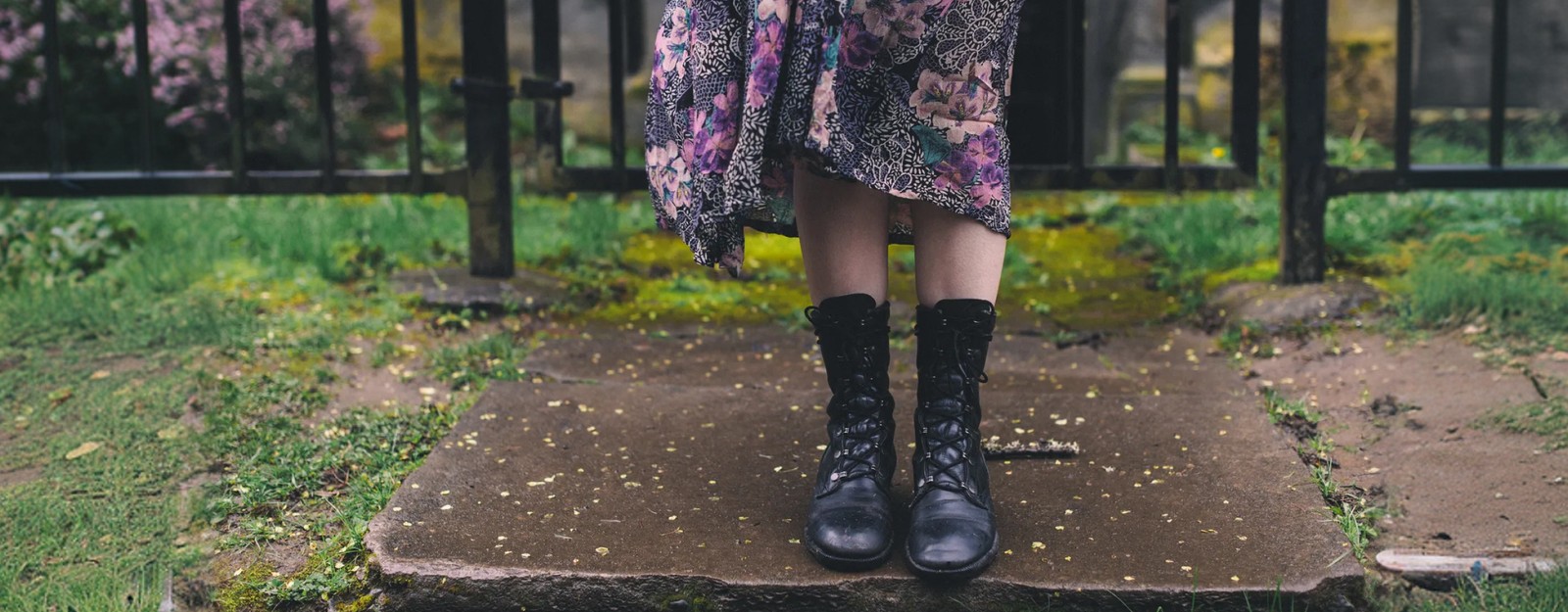 Aufnahme der Beine einer Person, die ein Kleid mit floralem Muster in Lila- und Rosatönen sowie schwarze Schnürstiefel trägt. Die Person steht auf einer Steinplatte in einem Garten; im Hintergrund sind grüner Rasen und ein schwarzer Metallzaun zu sehen.