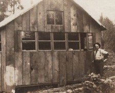 Hombre de pie junto a cabaña de madera rural escena de campamento de madera de la década de 1910