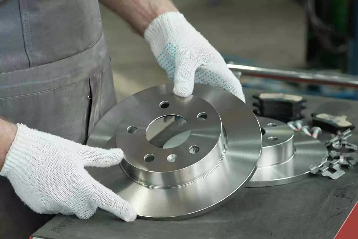 A mechanic with white gloves picks up a solid brake rotor. A second rotor is on the work table surface, along with brake pads and clips.
