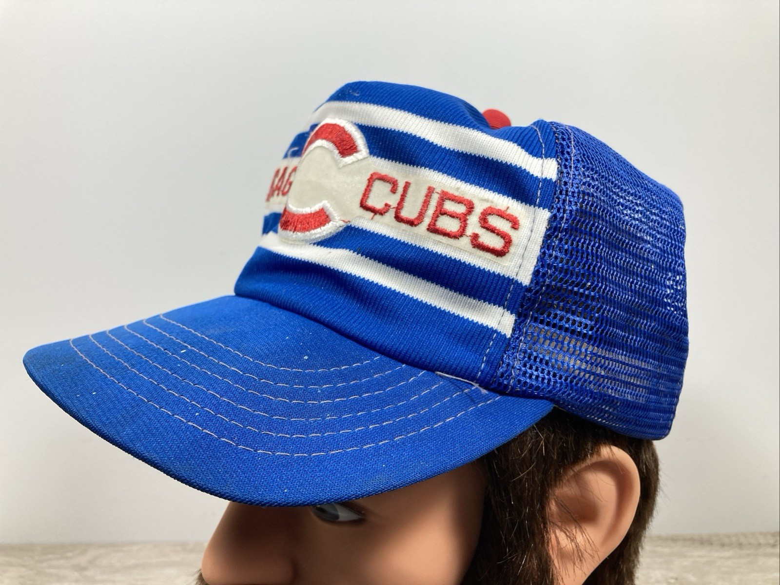 Blue snapback trucker hat with white and red stripe, embroidered “C” logo on front, and mesh fabric.