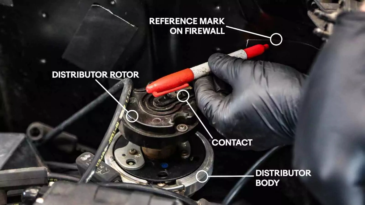 A mechanic places a reference mark on the firewall with a felt tip marker. Annotations call out the distributor body, rotor, and contact, along with the reference mark location.