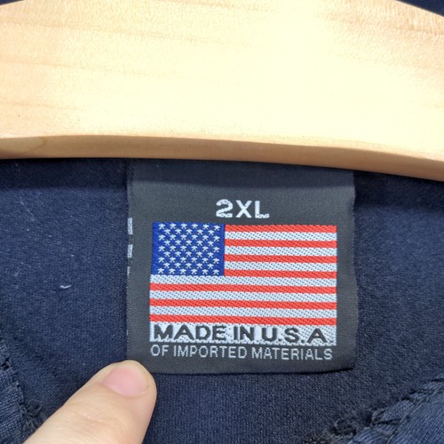 White fabric tag on blue “Made in U.S.A.” label, 2XL size, with American flag design and red, white, and gray text.